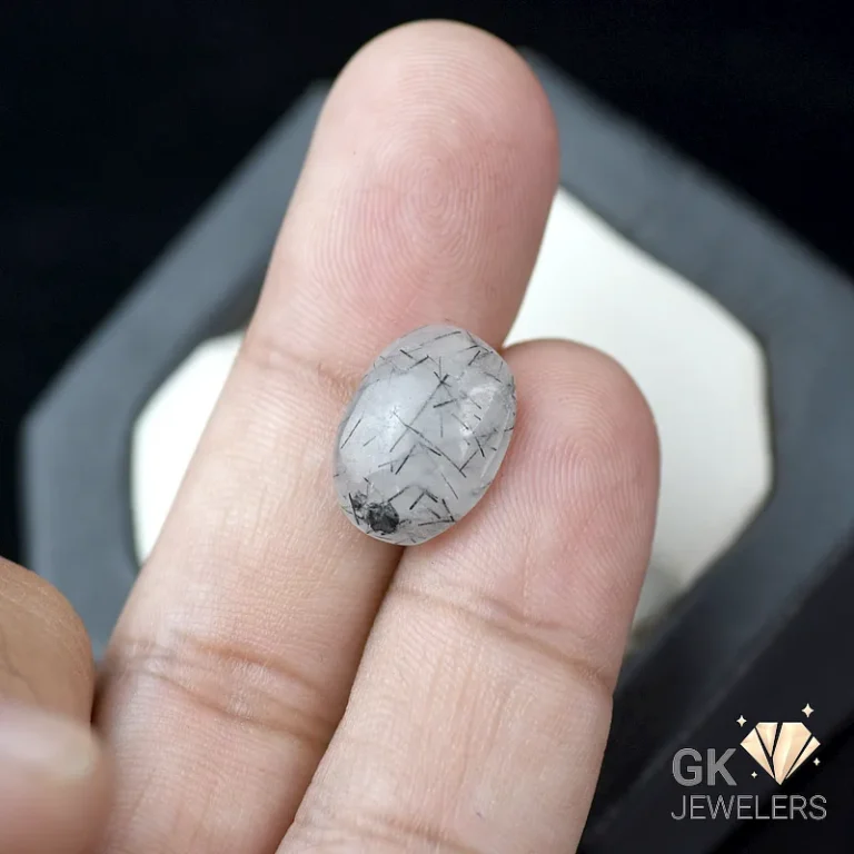 Oval white gemstone with black rutile inclusions held between fingers