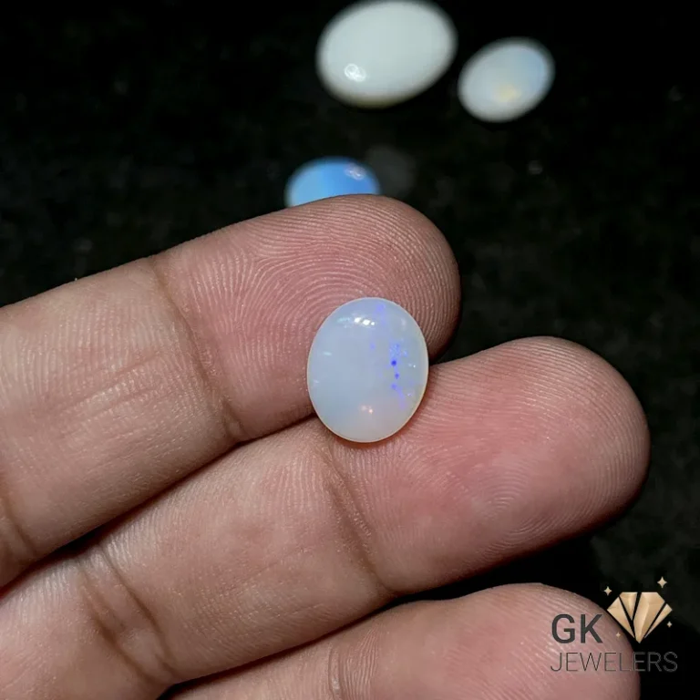Oval white opal gemstone held between fingers, displaying iridescent blue specks