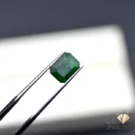 Emerald gemstone held by silver tweezers against a white background