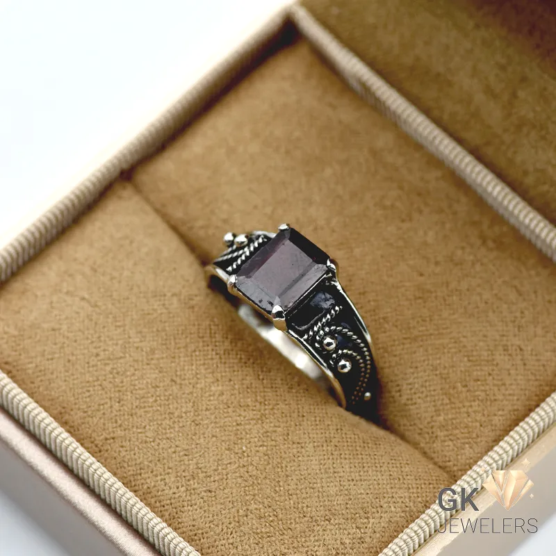 Purple garnet gemstone in emerald cut, set in handmade silver ring with decorative detailing, displayed in jewelry box