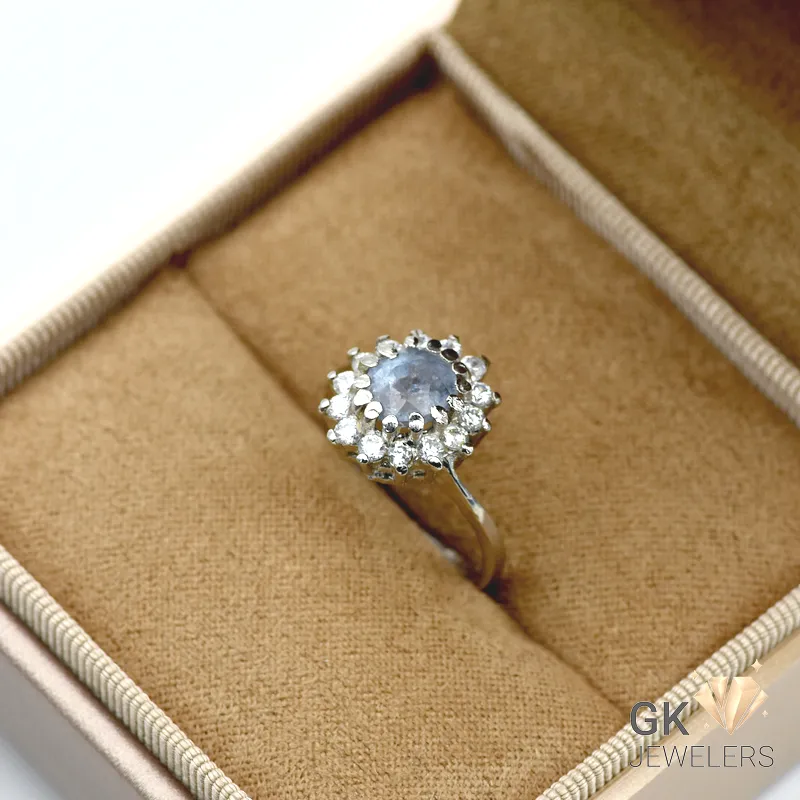 Round blue sapphire gemstone surrounded by clear stones in a silver ring setting, displayed in a beige jewelry box with 'GK JEWELERS' watermark