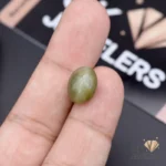 Hand holding a 7.75-carat green catseye gemstone between thumb and index finger, with a blurred 'GK JEWELERS' logo in the background