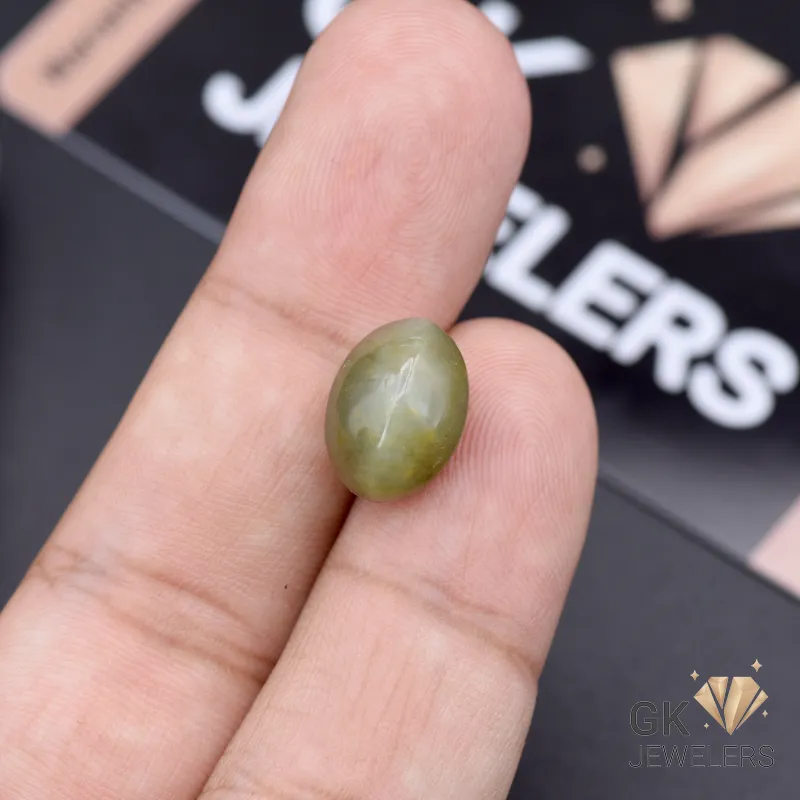 Hand holding a 7.75-carat green catseye gemstone between thumb and index finger, with a blurred 'GK JEWELERS' logo in the background