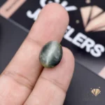 Hand holding an oval-shaped green catseye gemstone with chatoyant effect on a black background with 'GK JEWELS' logo