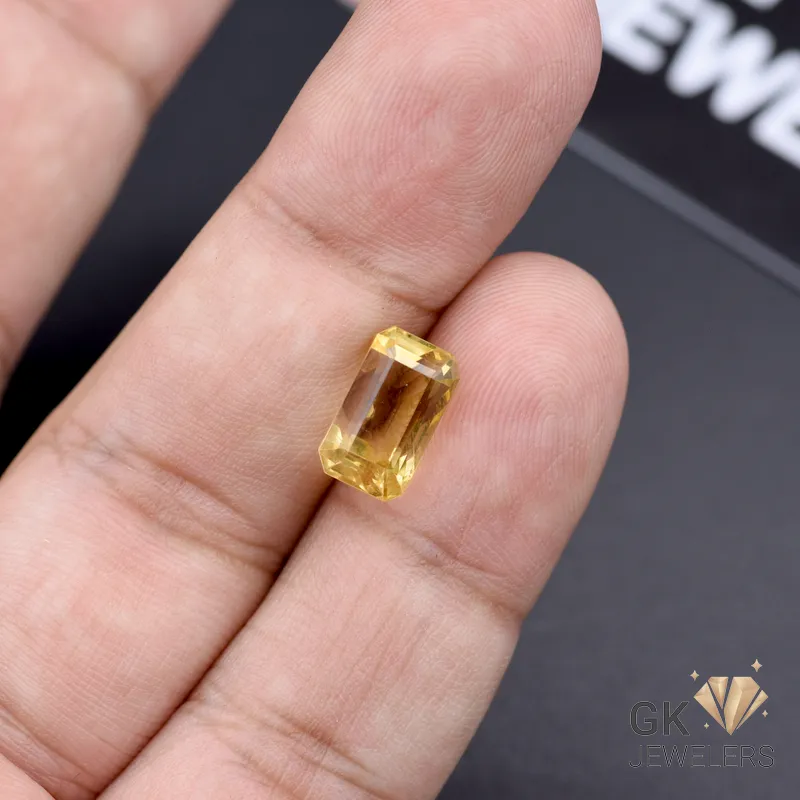 Emerald-cut yellow citrine gemstone held between fingers on a black background with 'GK JEWELS' logo