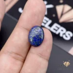 Oval-cut lapis lazuli gemstone held between fingers, displaying rich blue color with gold flecks