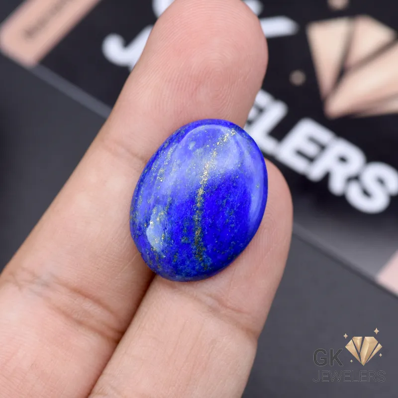 Hand holding an oval-cut natural Lapis Lazuli gemstone with golden pyrite inclusions, weighing 16.85 carats from Afghanistan