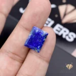 Emerald-cut lapislazuli gemstone held between fingers, showcasing deep blue color and characteristic opaque transparency