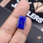 Hand holding a 9.55-carat emerald-cut lapis lazuli gemstone with vibrant blue color and gold flecks, showcasing its opaque transparency