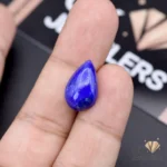 Hand holding a teardrop-shaped lapis lazuli gemstone with deep blue color and golden specks
