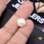 White natural pearl gemstone held between fingers on a dark surface with 'GK JEWELERS' logo