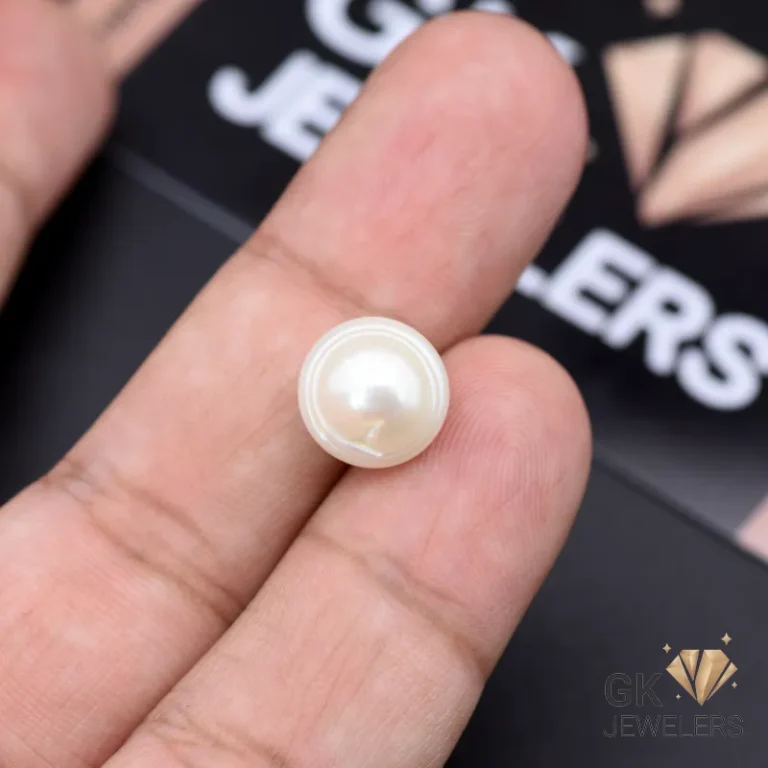 White natural pearl gemstone held between fingers on a dark surface with 'GK JEWELERS' logo