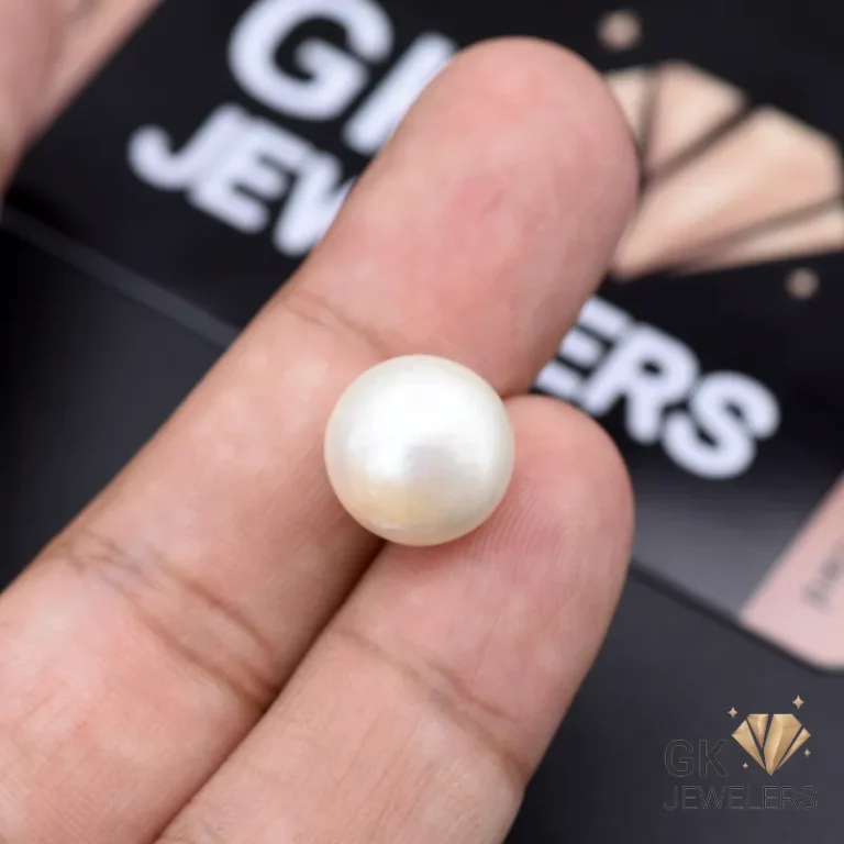Hand holding a large, round white pearl gemstone against a dark background with 'GK JEWELERS' branding