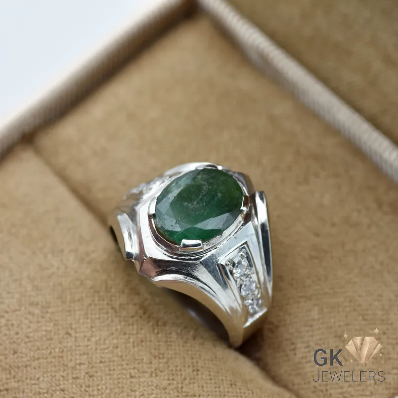 Silver ring featuring a large oval-cut green emerald gemstone with triangular diamond accents on either side, set in a handmade silver band, displayed on a beige fabric surface
