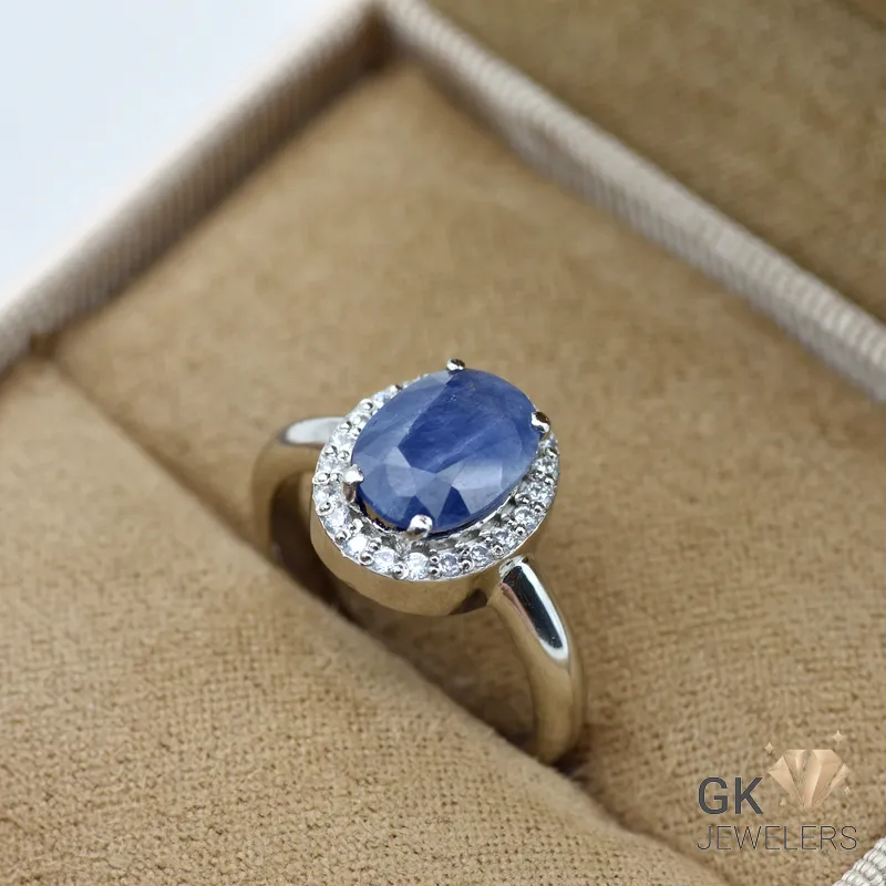 Oval blue sapphire gemstone surrounded by white diamonds in a silver ring setting, displayed on a beige jewelry box cushion