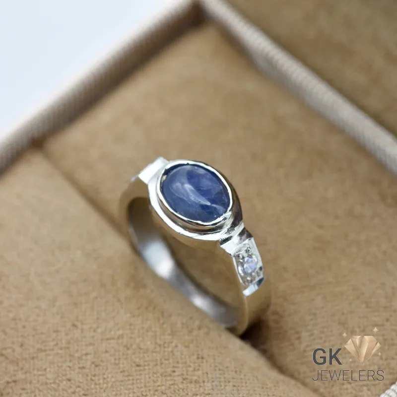 Oval blue sapphire gemstone set in sterling silver ring with diamond accents, displayed in a tan jewelry box