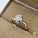 Oval-shaped white opal gemstone set in a handmade silver ring, showcasing iridescent colors, on a beige jewelry box fabric