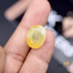 Finger holding a 19.29-carat oval-cut yellow sapphire gemstone with semi-transparent clarity