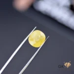 Faceted yellow sapphire gemstone held between silver tweezers on dark surface