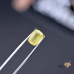 Cushion-cut yellow sapphire gemstone held by silver tweezers on a dark surface