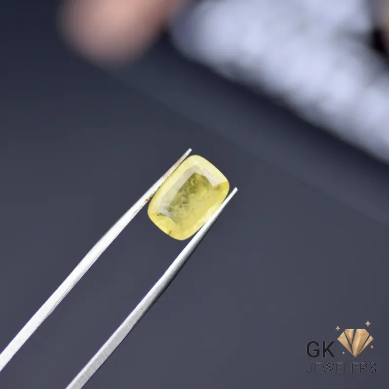 Cushion-cut yellow sapphire gemstone held by silver tweezers on a dark surface
