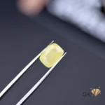 Cushion-cut yellow sapphire gemstone held by silver tweezers on dark surface