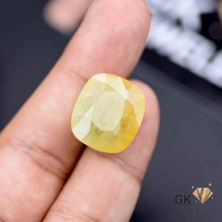 Cushion-cut 23.39-carat yellow sapphire gemstone held between fingers on a dark surface with blurred jewelry background