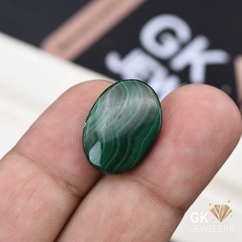Oval polished malachite gemstone held between person's fingers with banded patterns